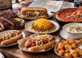 Table Filled With Regional American Foods Including Barbecue Ribs, Cheesesteak, Chili, Coney Dog, Clam Chowder And Deep-Dish Pizza.